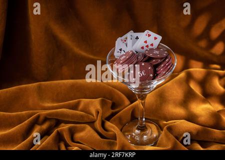 Rote Plastik-Poker-Chips liegen in einem Glas margarita-Glas auf einem braunen Samttuch. Ein Fan kleiner Spielkarten am Rand eines Glases. Stockfoto