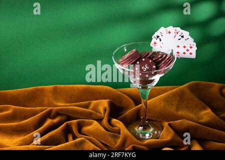 Rote Plastik-Poker-Chips liegen in einem Glas margarita-Glas auf einem braunen Samttuch. Ein Fan von kleinen Spielkarten am Rand eines Glases, wie ein Cocktail g Stockfoto