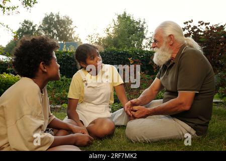 Zwei afroamerikanische Geschwister hören ihrem Großvater beim Plaudern zu, während sie vor ihm auf einem grünen Rasen in natürlicher Umgebung sitzen Stockfoto