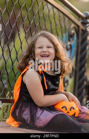 Ein süßes kleines Mädchen in einem Hexenkostüm für Halloween spaziert im Park in einem Korb mit Süßigkeiten in Form eines Kürbiss. Hochwertiges Foto Stockfoto