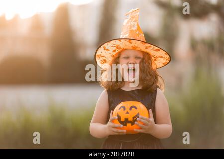 Ein süßes kleines Mädchen in einem Hexenkostüm für Halloween spaziert im Park in einem Korb mit Süßigkeiten in Form eines Kürbiss. Hochwertiges Foto Stockfoto