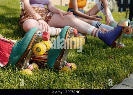 Rollschuhlaufen auf den Füßen einer jungen aktiven Frau, die auf grünem Gras sitzt und sich nach einer Freizeitaktivität im Park gegen ihre beiden Freunde entspannt Stockfoto