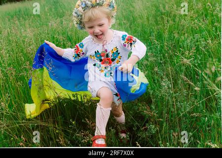 Die ukrainische Flagge ist auf den Schultern eines kleinen ukrainischen Mädchens. Glückliches ukrainisches Kind in freier Ukraine ohne Krieg läuft auf einem Feld entlang einer Landstraße Stockfoto