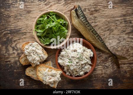 Eine Schüssel hausgemachter, geräucherter Makrelenpastete, serviert auf Brot mit einem Ruckelsalat und einer ganzen heißen geräucherten Makrele, Scomber scombrus, daneben. England Stockfoto