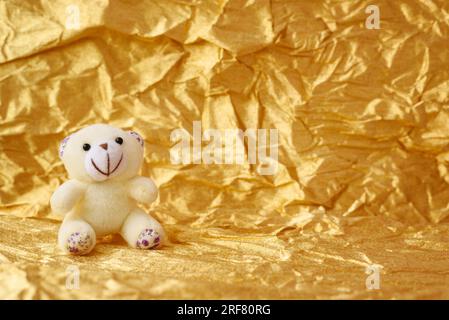 Der weiße Stoffbär Teddybär sitzt auf goldenem Papierhintergrund, Kopierbereich, Vorlage. Stockfoto
