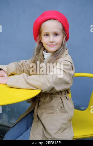Hübsches kleines Mädchen in einem Trenchcoat, rote Baskenmütze und Jeans posieren, während es am Tisch eines Straßencafés sitzt. Vertikales Foto Stockfoto