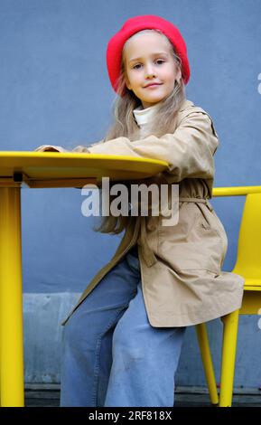 Hübsches kleines Mädchen in einem Trenchcoat, rote Baskenmütze und Jeans posieren, während es am Tisch eines Straßencafés sitzt. Vertikales Foto Stockfoto