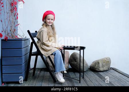 Ein hübsches kleines Mädchen in einem Trenchcoat, einer roten Baskenmütze und Jeans posiert, während es an einem Tisch in einem Straßencafé sitzt. Ein Mädchen, das mit dem Rücken zum Camer sitzt Stockfoto