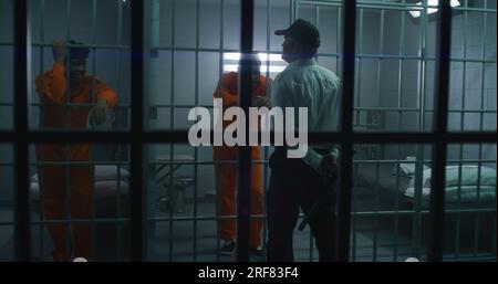 Ein Gefängniswärter mit Polizeistab steht vor Gefängniszellen und spricht mit Gefangenen. Afroamerikanische Kriminelle werden im Gefängnis inhaftiert. Mörder in der Strafvollzugsanstalt oder in der Haftanstalt. Stockfoto