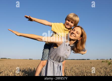 mutter und Sohn, 6-7 Jahre alt, spielen zusammen in der Natur. Eine Frau hält das Kind von oben auf dem Rücken, spreizt fröhlich die Arme und lacht. Fröhlicher Zeitvertreib auf Su Stockfoto