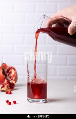 Ein Mann gießt Granatapfelsaft aus einer Flasche in ein großes Glas mit Eis. Das Konzept von Frische und Nützlichkeit. Im Hintergrund: Die Frucht und die Samen Stockfoto