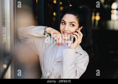 Attraktive Frau mit dunklen Haaren, ansprechenden Augen und vollen Lippen in eleganter Kleidung, die mit ihrer besten Freundin telefoniert, zur Seite schaut. Menschen, b Stockfoto