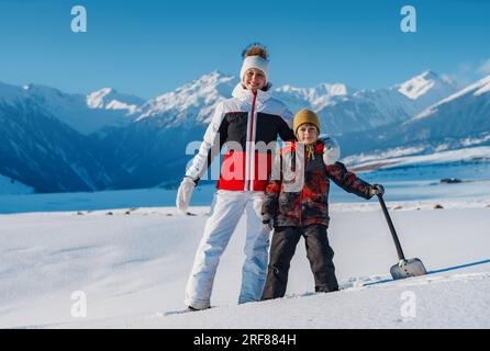 Frau und Kind mit Schaufel, die auf dem Hintergrund der Winterberge steht Stockfoto