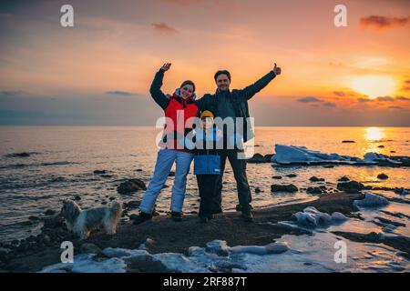 Vater, Mutter, Sohn und Hund glückliche Familie, die im Winter auf dem Sonnenuntergang am See posiert Stockfoto