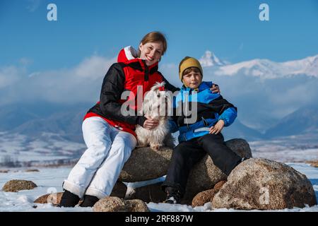 Mutter, Sohn und Shih-Tzu-Hund sitzen auf Steinen im Hintergrund der Winterberge Stockfoto