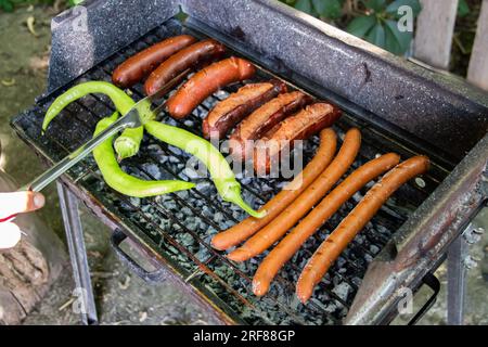 Holzkohlegrill Barbecue Grill im Hinterhof des Landes, bereitet Würste und grüne Paprika auf Grill mit Holzzaun im Hintergrund zu und grillt sie Stockfoto