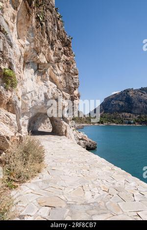 Fotografieren Sie das Arvanitia Walking Area, den Küstenweg in Nafplio, mit einem breiten gepflasterten Pfad, der zu einem Tunnel durch die Klippen, das Meer, den Strand und führt Stockfoto