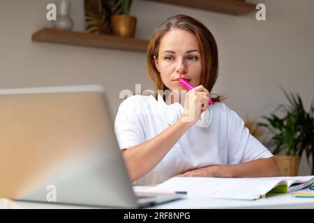 Junge Erwachsene Frau, die Hausaufgaben eines Online-Kurses mit het Laptop und Arbeitsbuch macht, Fernunterricht von zu Hause aus. Stockfoto