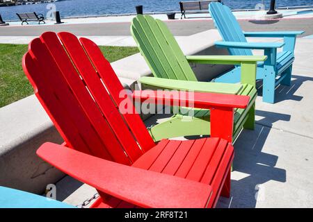 Eine Reihe leerer, farbenfroher Adirondack-Gartenstühle entlang der Hafenmauer Stockfoto