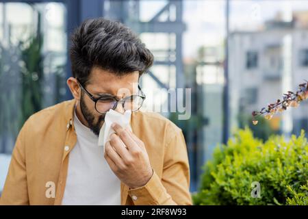 Ein junger Mann, der niest und eine Allergie gegen laufende Nase hat, sitzt tagsüber auf einer Bank vor einem Bürogebäude, ein hispanischer Geschäftsmann krank mit einem Gewebe in der Nähe seiner Nase. Stockfoto