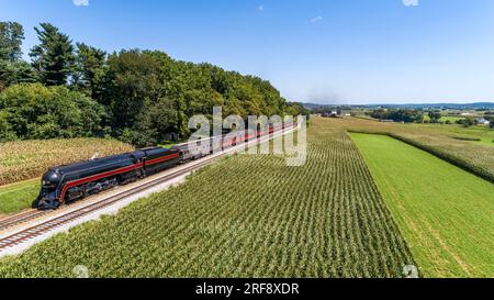 Ein Blick auf die Drohne eines antiken, stromlinienförmigen Dampflokomotivs, der an einem sonnigen Sommertag durch die Maisfelder fährt Stockfoto