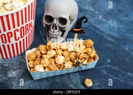 Pappschachtel mit leckerem Karamell-Popcorn und Schädel auf blauem Hintergrund, Nahaufnahme. Halloween-Feier Stockfoto