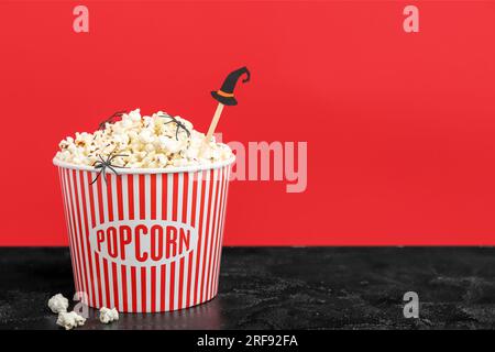 Eimer mit leckerem Popcorn mit Papierhut und Spinnen auf farbigem Hintergrund. Halloween-Feier Stockfoto