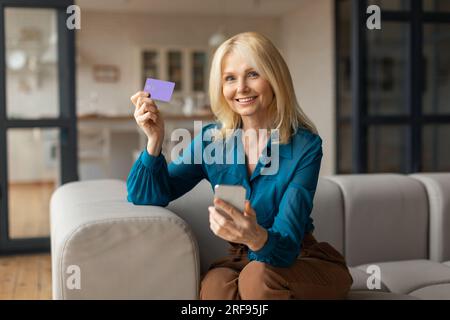 Mobiles Online-Shopping. Glückliche, reife Frau mit Kreditkarte und Handy, die auf dem Sofa saß und die Internetbestellung bestätigte Stockfoto