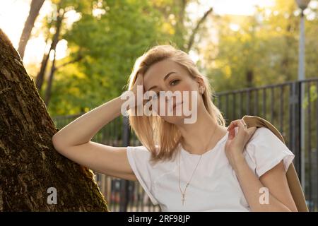 Müde Weiße echte Frau lehnt sich an den Baum und schaut in der Sommerzeit bei Sonnenuntergang auf die Kamera. Erholung und Erholung in Natur, Selbstwahrnehmung, Freizeit, geistig Stockfoto