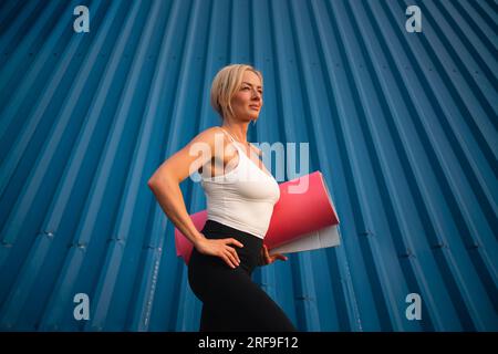 Porträt einer mittelalterlichen wunderschönen blonden Frau mit Sportmatte auf blauem Hintergrund. Gesunder, aktiver Lebensstil, Yoga-Workout Stockfoto