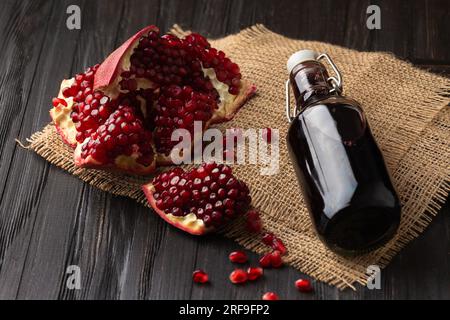 Reifer Granatapfelbruch, verstreute große Körner, frisch gepresster Granatapfelsaft in einer transparenten Flasche auf Holzhintergrund. Stockfoto