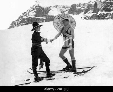 Vereinigte Staaten: c. 1923. Eine Skifahrerin am Berghang, die eine Schärpe und einen Westernhut trägt, schüttelt sich mit einem Mann auf Skiern in einem einteiligen Badeanzug und mit einem Sonnenschirm die Hände. Stockfoto