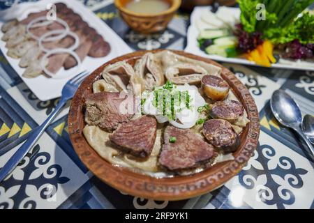 Traditionelles kasachisches asiatisches Gericht Beshbarmak: Nudeln mit Lamm und Zwiebeln, Nahaufnahme auf einem Teller auf dem Tisch. In der Nähe der Brühe in Schüsseln. dastarkhan. Bolied Stockfoto