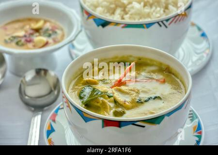 Eine Mahlzeit mit grünem thailändischem Curry-Huhn, serviert mit Reis in gemusterten Schüsseln im westlichen Stil auf einer weißen Tischdecke. Geringe Schärfentiefe. Stockfoto