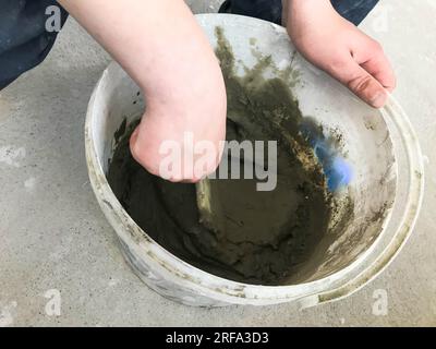 Baumeister-Hände mit einem Metallspachtel kneten in einem großen weißen Plastikeimer, Pflaster, Fliesenkleber, Zement für die Reparatur einer Wohnung, Hous Stockfoto