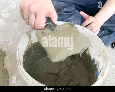 Baumeister-Hände mit einem Metallspachtel kneten in einem großen weißen Plastikeimer, Pflaster, Fliesenkleber, Zement für die Reparatur einer Wohnung, Hous Stockfoto