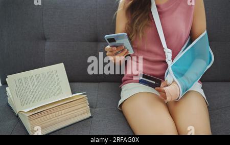 Junge asiatische Frau, die aufgrund von Wunde Arme eine weiche Schiene trägt, mit Telefon und Kreditkarte auf dem Sofa zu Hause. Stockfoto