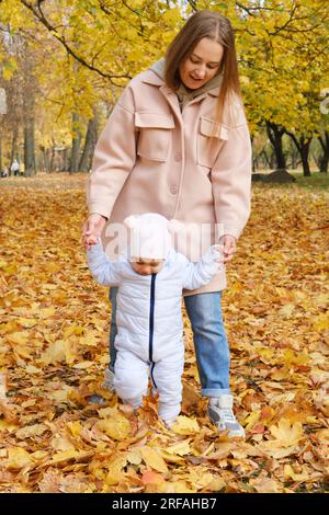 Mutter verbringt Zeit mit ihrer Tochter im Herbstpark. Mutter unterstützt ihre Tochter an den Händen und hilft ihr beim Gehen. Vertikales Foto Stockfoto