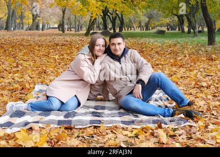 Junges Paar, Mann und Frau, sitzen auf einer Decke in einem Herbstpark, lächeln und in die Kamera schauen. Horizontales Foto Stockfoto