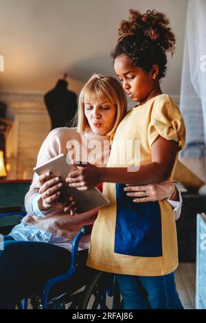 Glückliche Mutter im Rollstuhl mit Kind, das zu Hause ein digitales Tablet benutzt. Menschen Familie Glück Konzept. Stockfoto