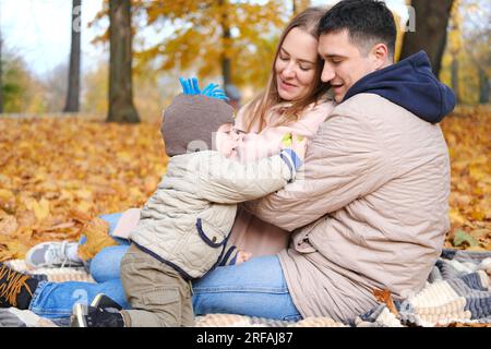 Familienwochenende. Junge Eltern mit einem kleinen Sohn verbringen Zeit zusammen im Herbstpark und sitzen auf einer Decke. Das Kind isst einen Apfel. Horizontales Foto Stockfoto