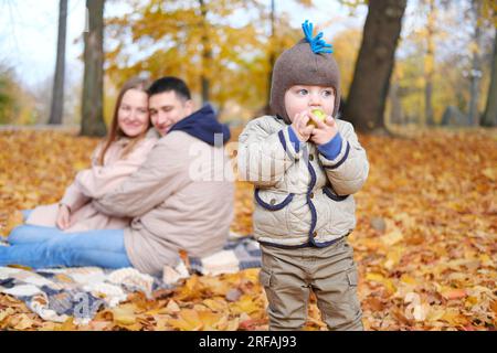 Familienwochenende. Junge Eltern und ihr kleiner Sohn verbringen Zeit zusammen im Herbstpark. Das Kind isst einen Apfel, die Eltern schauen ihn an und lächeln Stockfoto