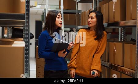 Weibliche Mitarbeiterin in Krücken, die Qualitätskontrolle mit Kleinunternehmer in der Nähe von Lagerregalen durchführt. Junge Frau mit körperlichen Problemen, die mit einem Laptop für die Supply-Chain-Produktion arbeitet. Stockfoto