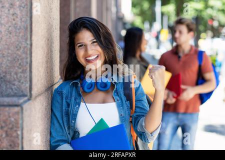 Weibliche Studentin mit Abschluss zufrieden mit der Prüfung mit Freunden im Hintergrund vor dem Universitätsgebäude im Sommer in der Stadt Stockfoto