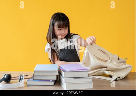 Ein süßes, junges asiatisches Mädchen packt ihre Schultasche und legt ihre Bücher in einen Rucksack, bereitet sich auf den ersten Schultag vor, auf einem isolierten gelben Ba Stockfoto