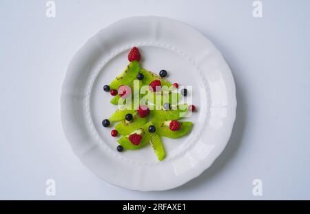 weihnachtsessen für Kinder. Weißes Gericht mit weihnachtsbaum aus Kiwi, Himbeeren, Cowberry und huckleberry. Hochwertiges Foto Stockfoto