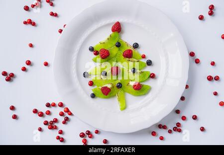 weihnachtsessen für Kinder. Weißes Gericht mit weihnachtsbaum aus Kiwi, Himbeeren, Cowberry und huckleberry. Hochwertiges Foto Stockfoto