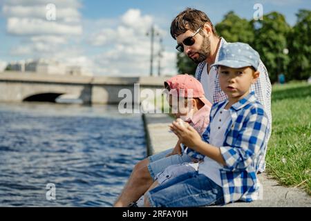 Ather und zwei Jungs, die am Ufer des Flusses Newa sitzen und Zeit miteinander verbringen, in Sankt petersburg, Russland. Bild mit selektivem Fokus. Stockfoto