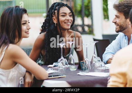 Eine vielfältige Gruppe von Millennials-Freunden, geführt von einer fröhlichen afroamerikanischen Frau, die eine gesellige Mahlzeit in einem Restaurant, ihr Lächeln und Lachen genießen Stockfoto