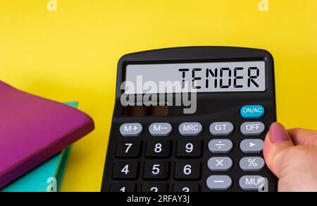 Das Wort „Tender“ auf einem Taschenrechner wird von einer Hand auf gelbem Hintergrund gehalten, mit Schreibwaren, Draufsicht Stockfoto